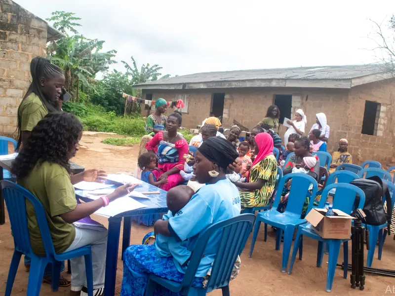 Women seated in rows outside a building while a volunteer registers them at a table.