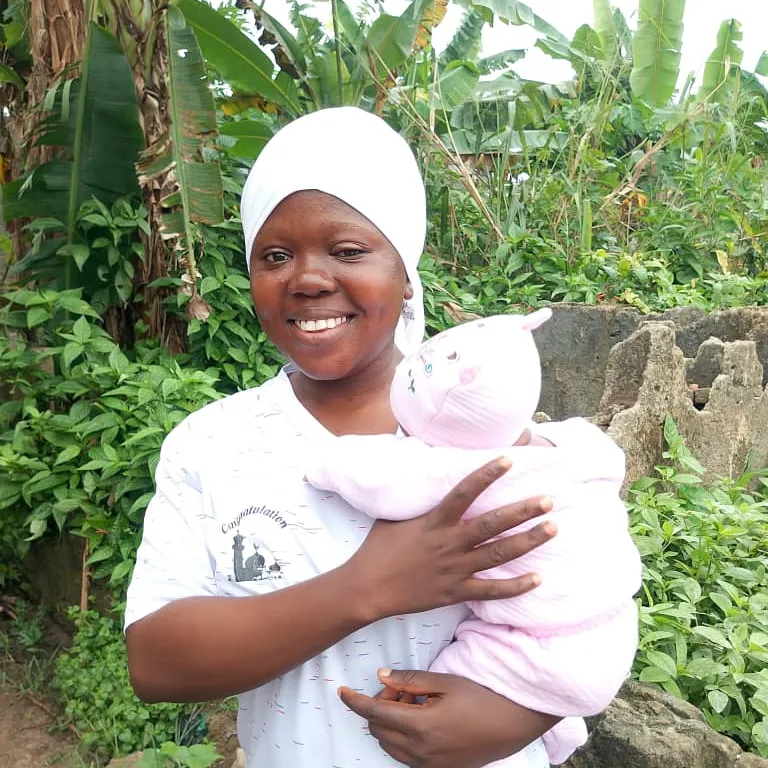 A smiling mother in a white head wrap holding her newborn, who is wrapped in pink, outdoors among green plants.